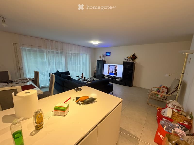 A modern living room with a blue sofa, coffee table, dining area, and television. The room features large windows with sheer curtains and a tiled floor.