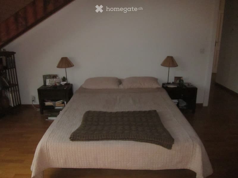 Cozy bedroom with wooden floor, slanted ceiling, bed with pillows and blanket, nightstands with lamps, books, and picture frames.