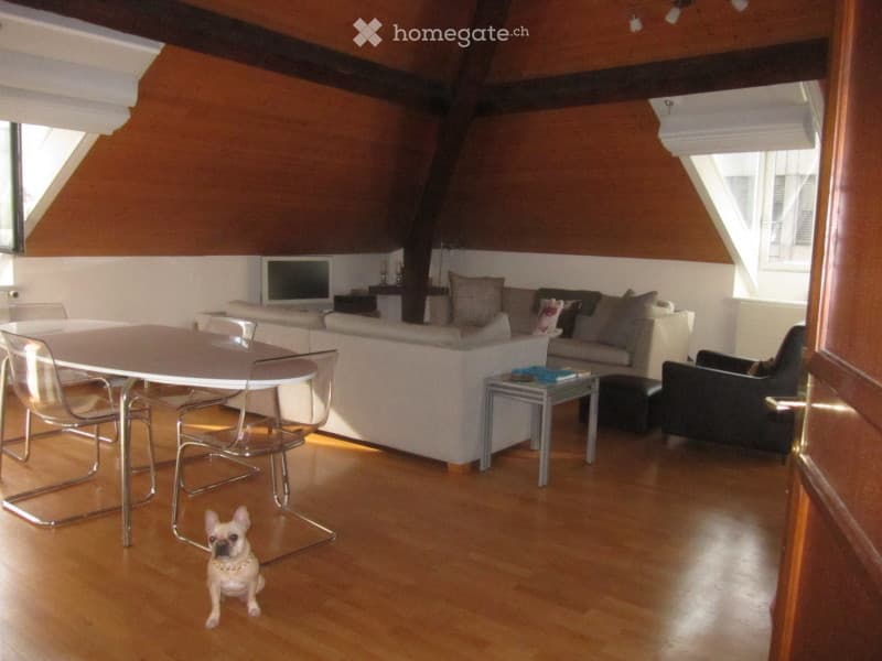 Bright living room with wooden floors, white walls, and exposed wooden beams. Features a white sofa, a small dining table with chairs, a television, and a small armchair.