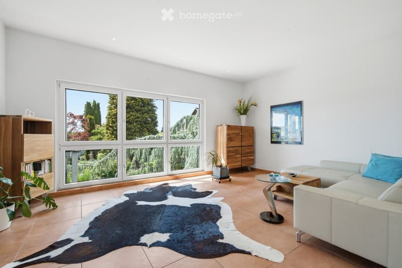 Bright living room with large windows overlooking a garden, featuring a white sofa, coffee table, wooden cabinet, and a cowhide rug.
