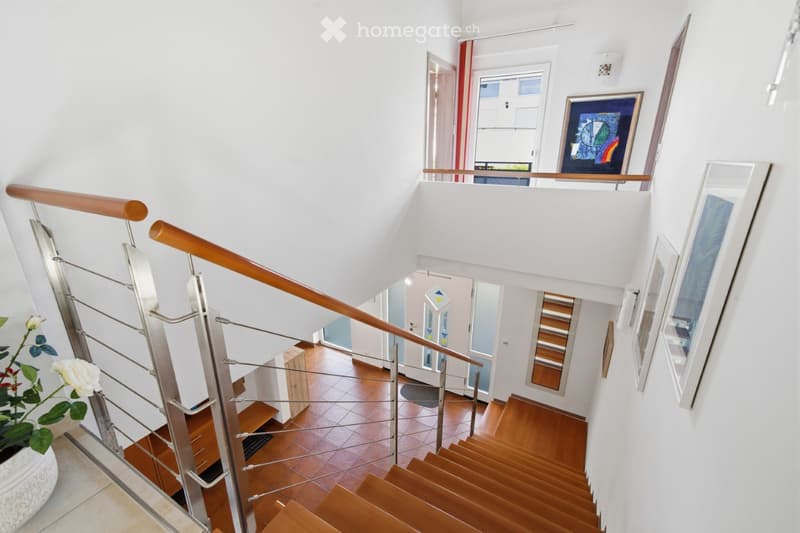 Modern interior staircase with wooden steps, metal railing, and white walls. The hallway features framed artwork and a potted plant on the left side.