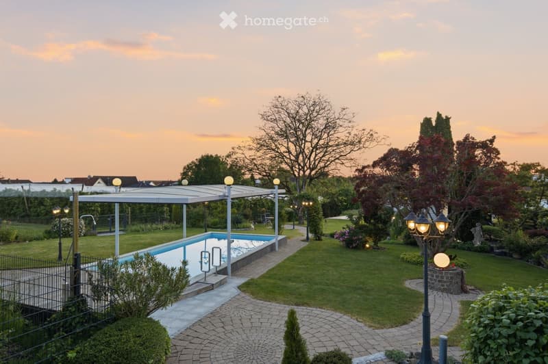 Well-maintained garden with a swimming pool, covered patio, neatly trimmed lawn, trees, shrubs, and decorative lighting.