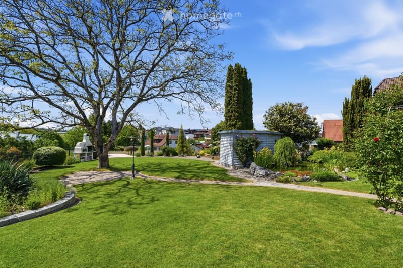 Well-maintained garden with a large tree, neatly trimmed lawn, pathways, and a small shed. The area includes a barbecue and surrounding greenery.