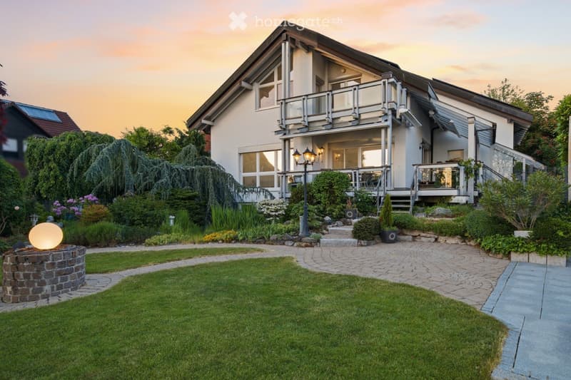 Two-story house with a modern design, featuring large windows, a balcony, and a well-maintained garden with a circular fire pit and decorative lighting.