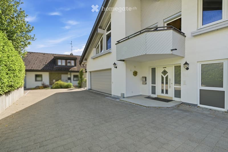 Two-story house with white exterior, garage, and balcony on the second floor. The driveway is paved, and there is a well-maintained garden on the left side.