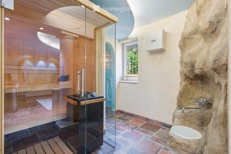 Bathroom with a sauna area, stone sink, toilet, and bathtub, featuring a large window and a decorative stone wall.