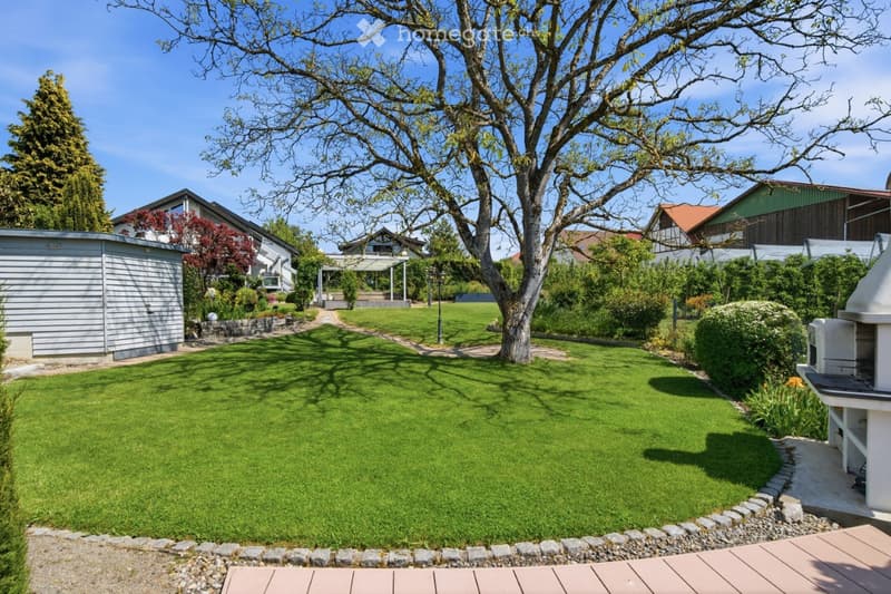 Well-maintained garden with a large tree, neatly trimmed lawn, surrounding shrubs, and a paved pathway leading to a covered patio area.
