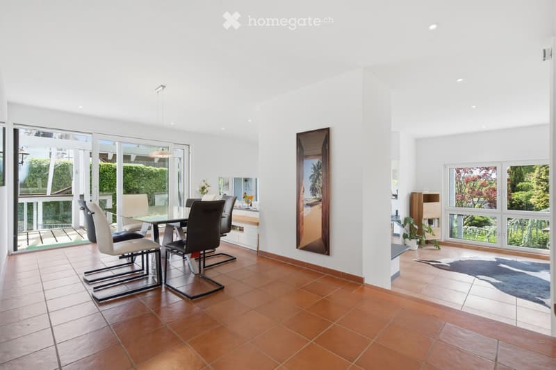 Bright dining area with a glass dining table, modern chairs, a large mirror on the wall, and a potted plant near the window. The space features large windows and a view of greenery outside.