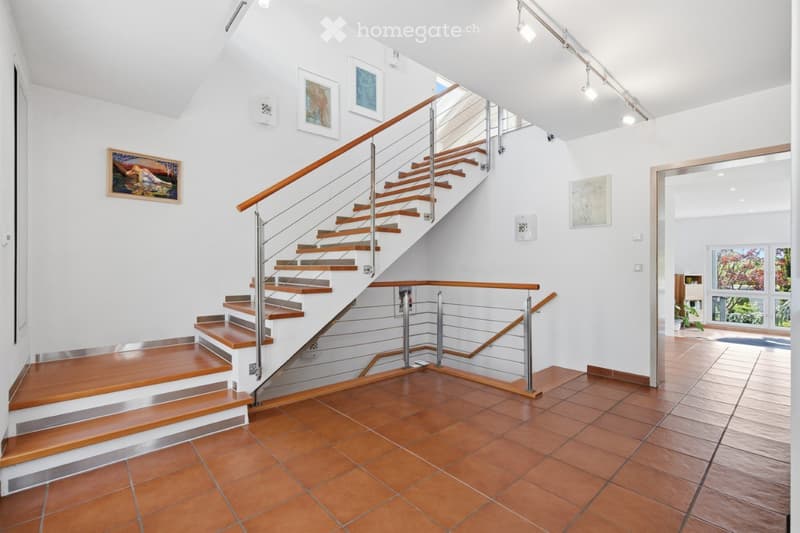 Modern hallway with wooden staircase, metal railing, and framed artwork on the walls. The floor is tiled in warm terracotta, and the ceiling features recessed lighting.