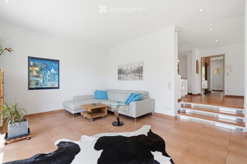 Modern living room with white walls, tiled floor, white sectional sofa with blue cushions, wooden coffee table, glass side table, cowhide rug, and artwork on the walls.
