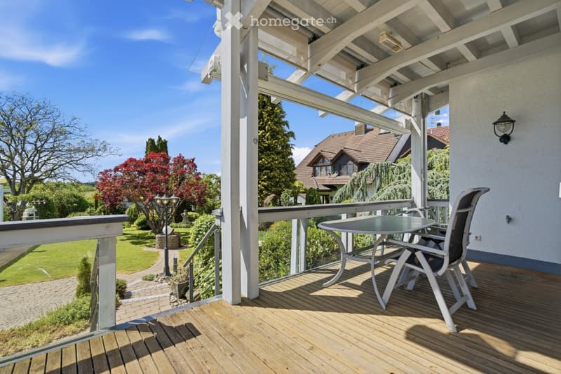 Wooden balcony with a table and chairs, overlooking a landscaped garden with trees and a house in the background.