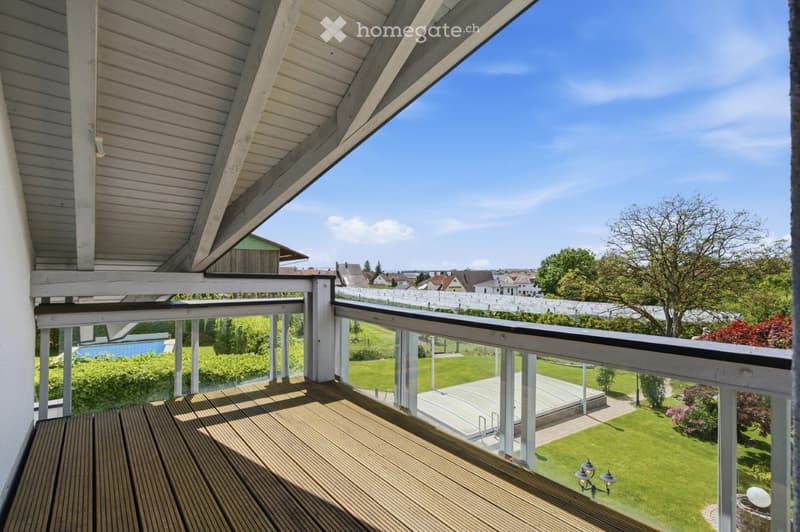 Wooden balcony with a glass railing, overlooking a well-maintained lawn and garden area with trees and a small structure in the distance.