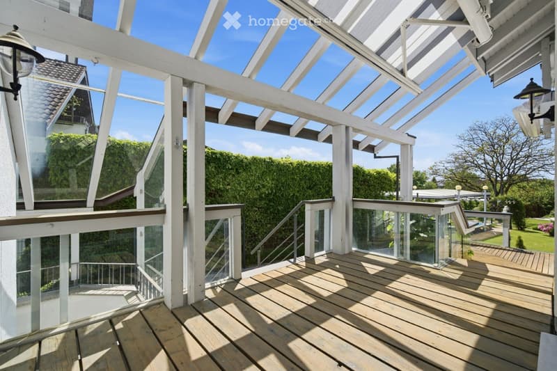 Wooden deck with glass railing, white wooden pergola structure, and outdoor lighting fixtures.