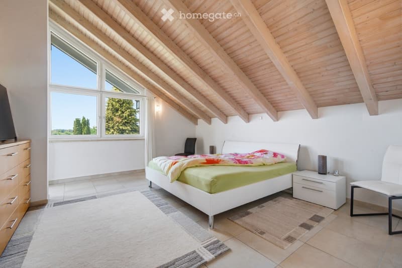 Bright bedroom with wooden slanted ceiling, large windows, white walls, tiled floor, and modern furniture including a bed, nightstand, chair, dresser, and lamp.