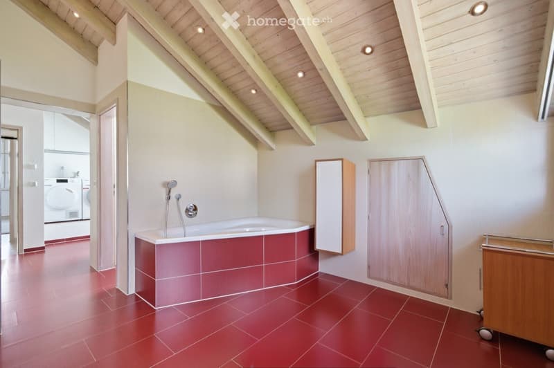 Modern bathroom with red tiled floor, white bathtub, wooden cabinets, and a visible washing machine in the adjacent space.