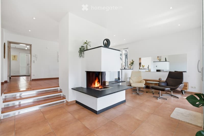 Modern living room with a central fireplace, two armchairs, a coffee table, and a small table near the fireplace. The room features wooden flooring and white walls.