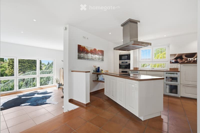 Bright, modern kitchen with white cabinets, a central island, stainless steel appliances, and large windows overlooking a green landscape.