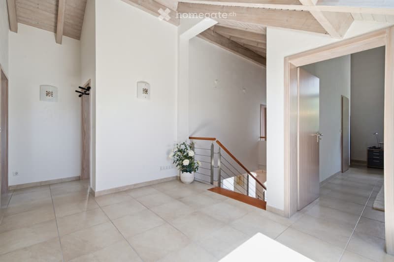 Bright hallway with tiled flooring, white walls, wooden ceiling beams, and a staircase leading upwards. Features a small potted plant on the floor and several doors leading to other rooms.