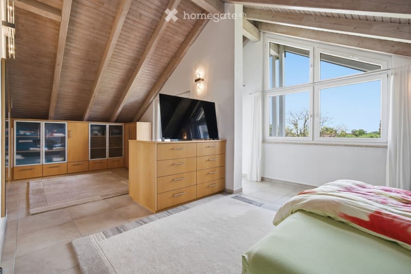 Bright bedroom with wooden slanted ceiling, large windows, bed with colorful bedding, TV on a wooden stand, and built-in wooden cabinetry.