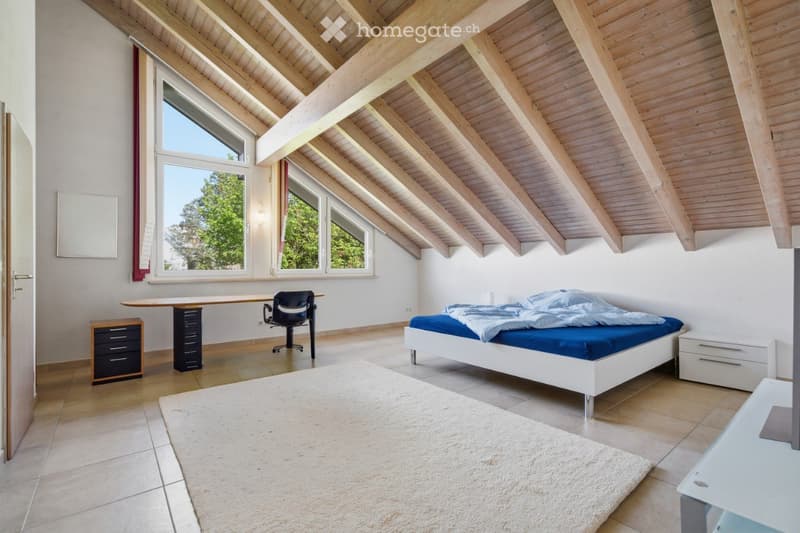 Bright bedroom with wooden slanted ceiling, large windows, bed with blue bedding, desk with chair, nightstands, and a rug.
