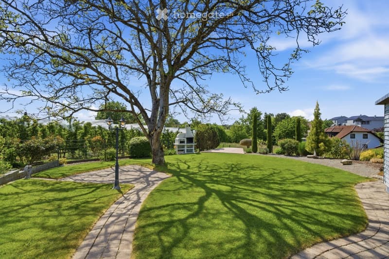 Well-maintained garden with a large tree, neatly trimmed lawn, stone pathways, and a small outdoor fireplace structure.