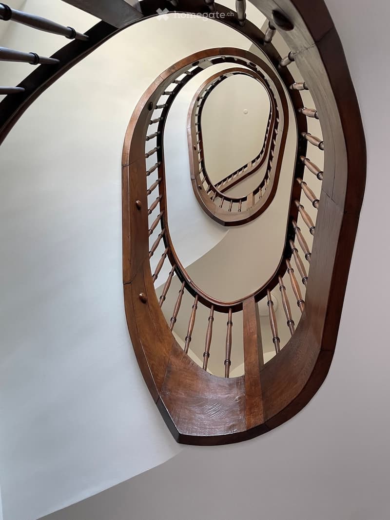 Spiral staircase with wooden handrails and balusters, white walls, and a polished wooden floor.