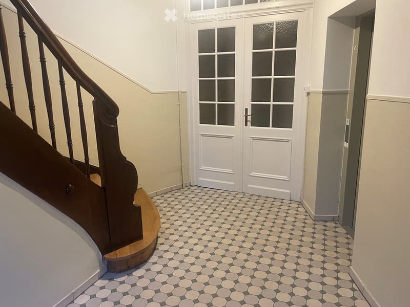 Hallway with a staircase featuring a wooden handrail and a white door with glass panels at the end. The floor is covered with hexagonal tiles in shades of gray and beige.