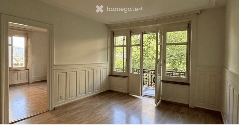 Empty room with wooden flooring, large windows, and a balcony access. Walls are painted in a light color with decorative paneling.
