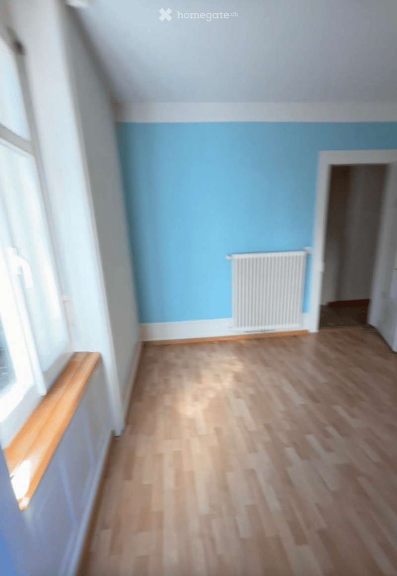 Empty room with light blue walls and wooden flooring, featuring a radiator and a doorway leading to another space.