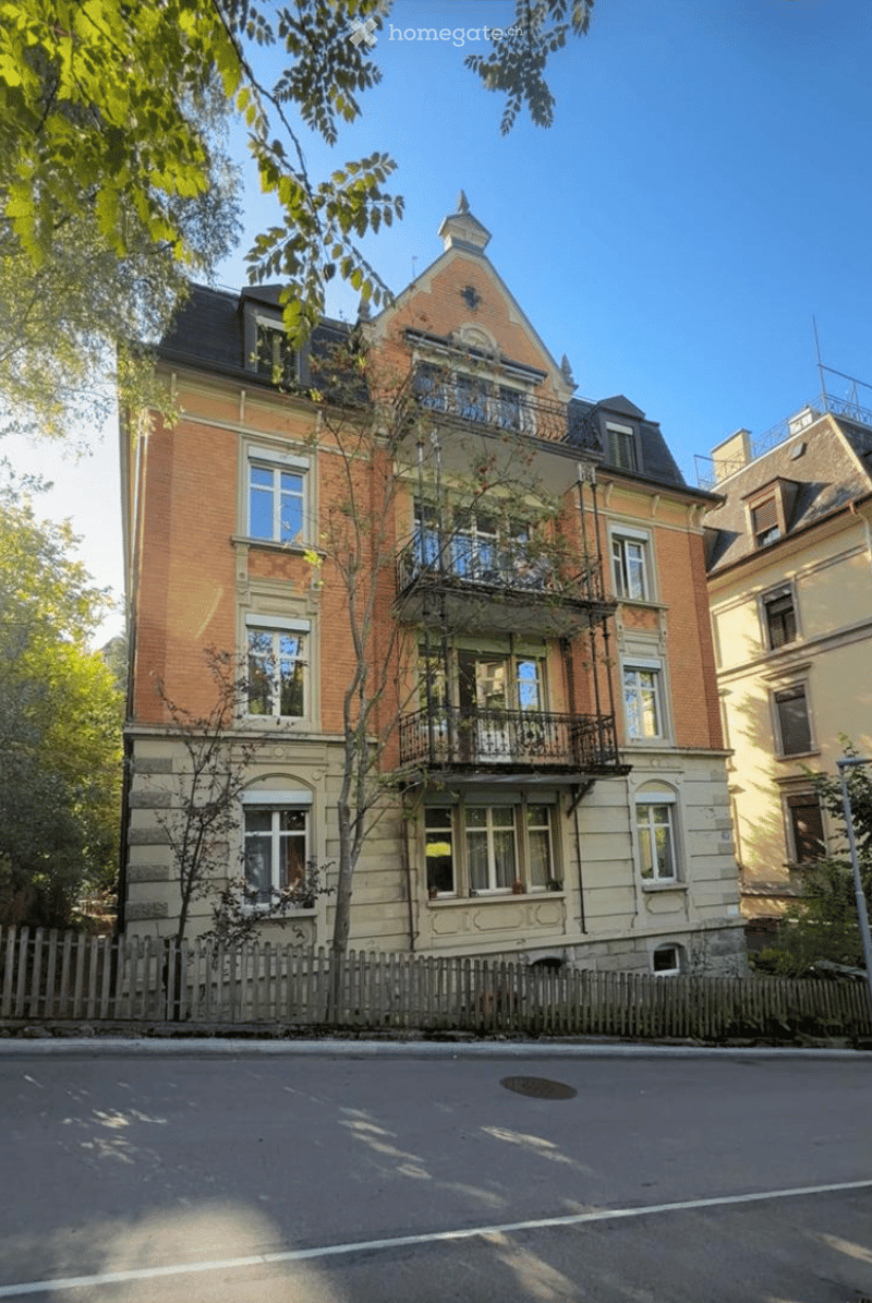 A multi-story brick building with a balcony and ornate architectural details, surrounded by trees and a wooden fence.
