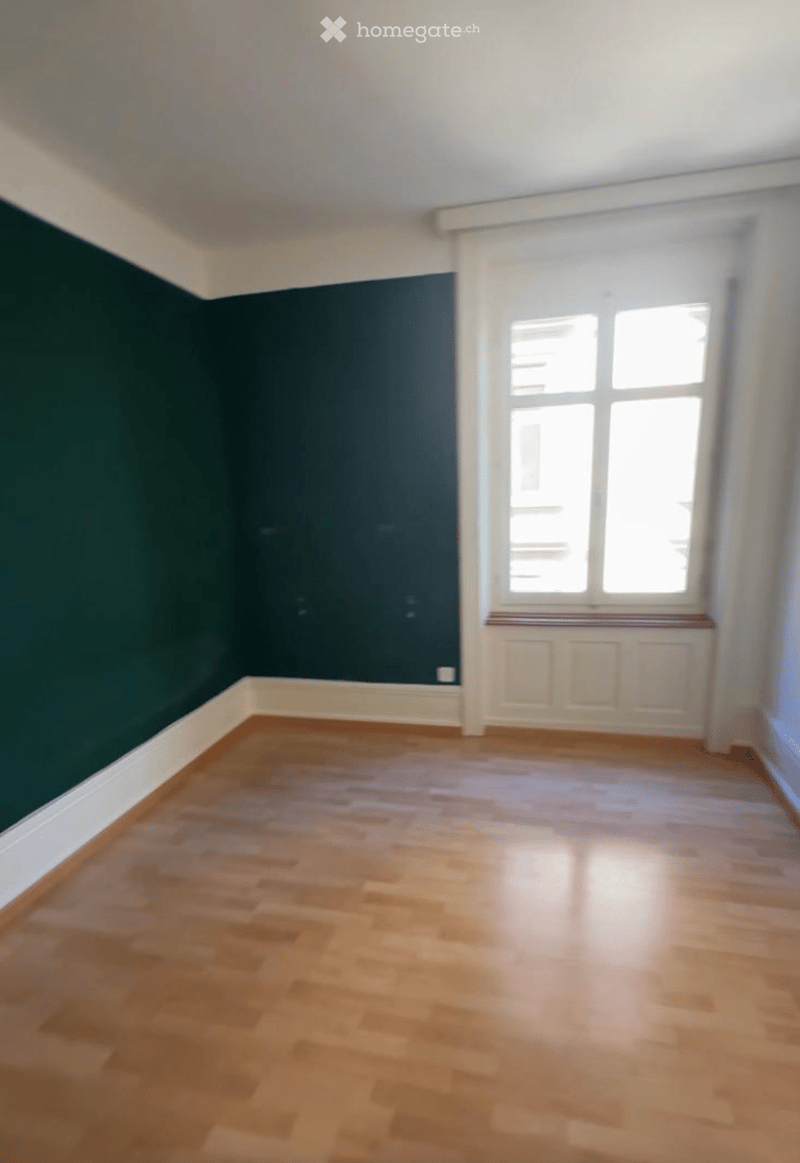 Empty room with wooden flooring, dark green accent wall, white trim, and large windows with white frames.