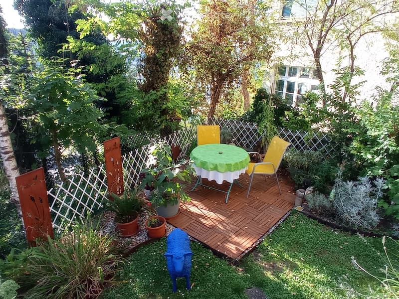 Backyard garden with patio, wooden deck, table, chairs, various plants, shrubs, decorative wood sculptures, and a blue animal statue.