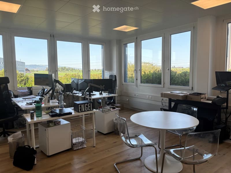 Office space with desks, chairs, laptops, and a round meeting table with chairs. Large windows offer a scenic view of the countryside.