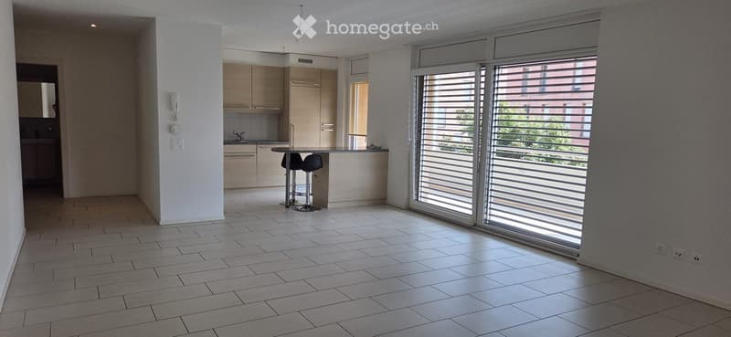 Bright, open-plan living and dining area with tiled flooring, large sliding glass doors leading to a balcony, and a small kitchen area with cabinets and a sink.