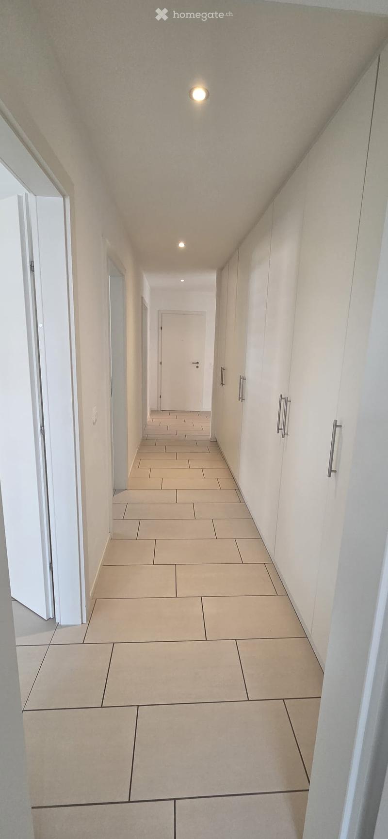 Clean, modern hallway with beige tiled flooring, white walls, and recessed lighting. Features built-in white cabinets with silver handles on one side and several doors leading to other rooms.
