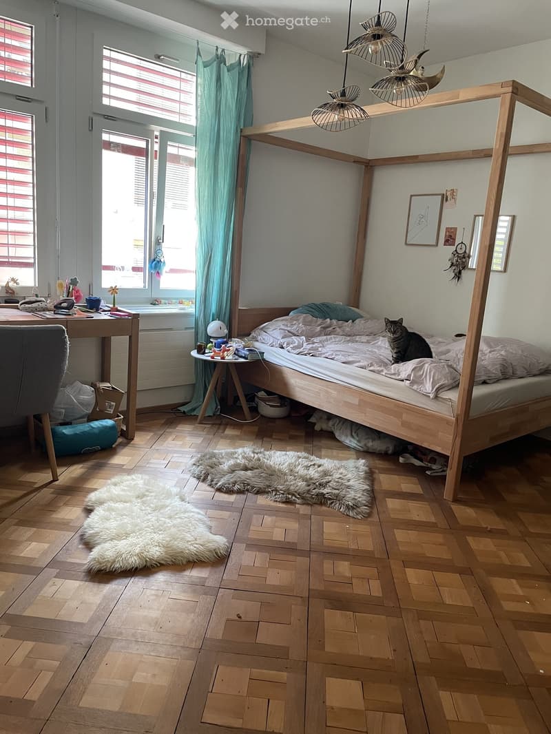 Cozy bedroom with wooden floor, lofted bed, desk, and two furry rugs. Features a cat on the bed, a small table with items, and modern lighting.