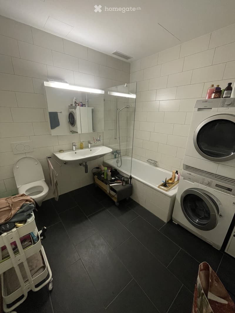 Modern bathroom with a toilet, sink, bathtub, and washing machine. Features a large mirror, tiled walls, and a small shelf with toiletries.