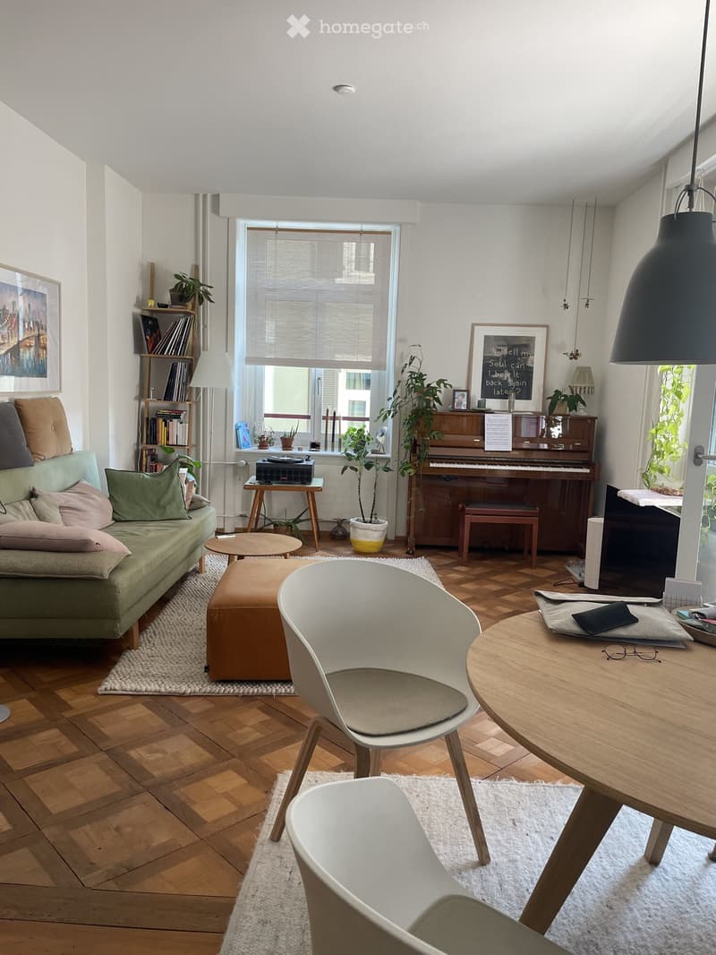 Living room with wooden flooring, large window, green sofa, ottoman, white chairs, round table, piano, bookshelf, and decorative plants.