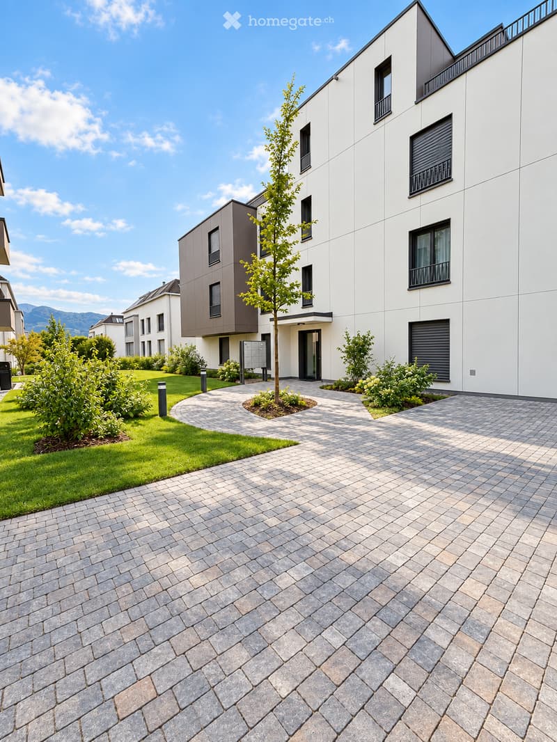 Modern residential building with white and grey facade, multiple windows, and a landscaped entrance area with a paved pathway and small garden.