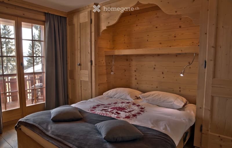 Cozy wooden bedroom with a large bed, pillows, and a nightstand lamp. Large windows offer a scenic view of the outdoors.