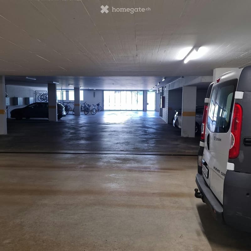 Underground parking area with concrete floors and pillars, several parked cars and bicycles, natural light from large windows at the far end.