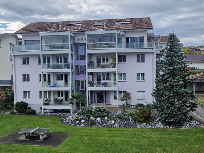 A modern, multi-story residential building with light purple walls, large windows, and multiple balconies. The building features a well-maintained lawn, a picnic table, and surrounding trees.