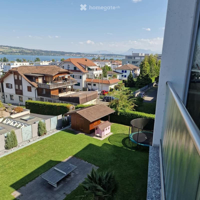 Well-maintained garden with green lawn, trampoline, small playhouse, and outdoor seating area. Surrounded by residential houses and a scenic lake view in the background.