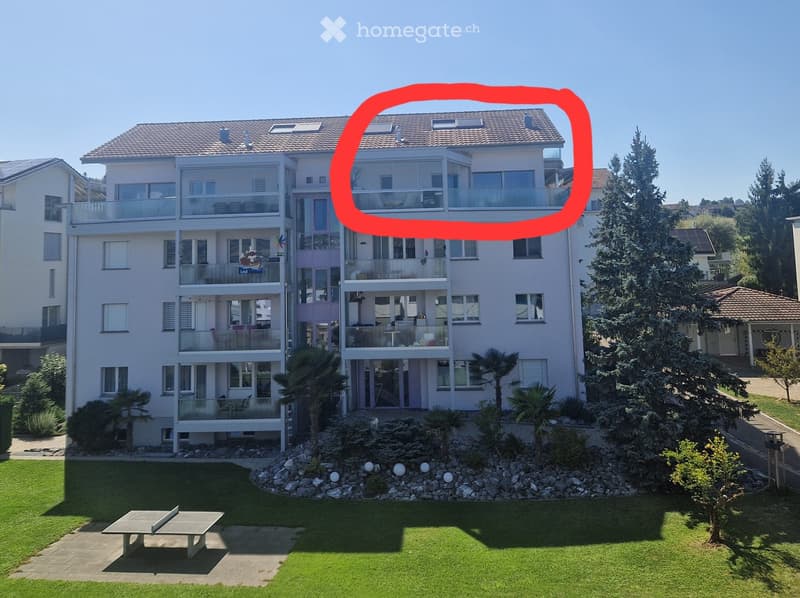 Modern multi-story residential building with balconies, surrounded by landscaped garden with trees, shrubs, and a small seating area.