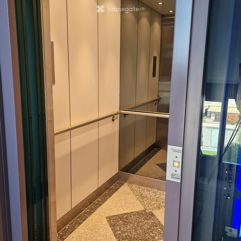 Modern hallway with light-colored walls, polished marble flooring, and a glass elevator door with a control panel visible.