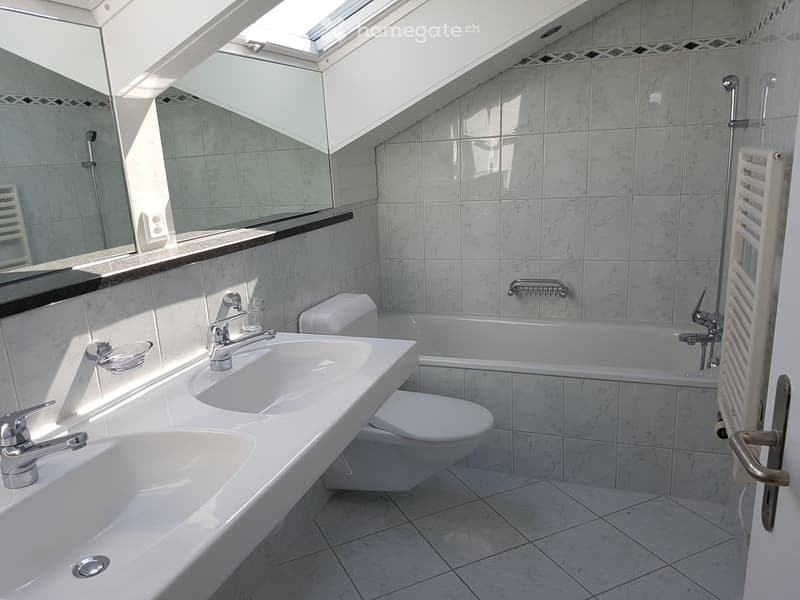 Modern bathroom with two sinks, a toilet, a bathtub, and a large mirror. The room features marble tiles and a skylight window providing natural light.