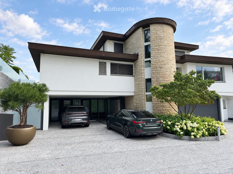 Modern two-story house with white and brown exterior, large windows, and a curved roofline. Two luxury cars are parked in the driveway, surrounded by landscaped greenery and a large planter.
