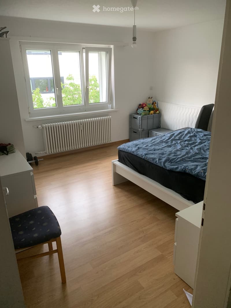 Bright bedroom with wooden flooring, large windows, a bed with blue bedding, a chair, a stool, and a radiator under the windows.