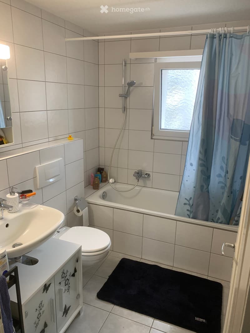 Modern bathroom with white tiles, a bathtub with a shower curtain, a sink with a mirror, a toilet, and a black bath mat on the floor.