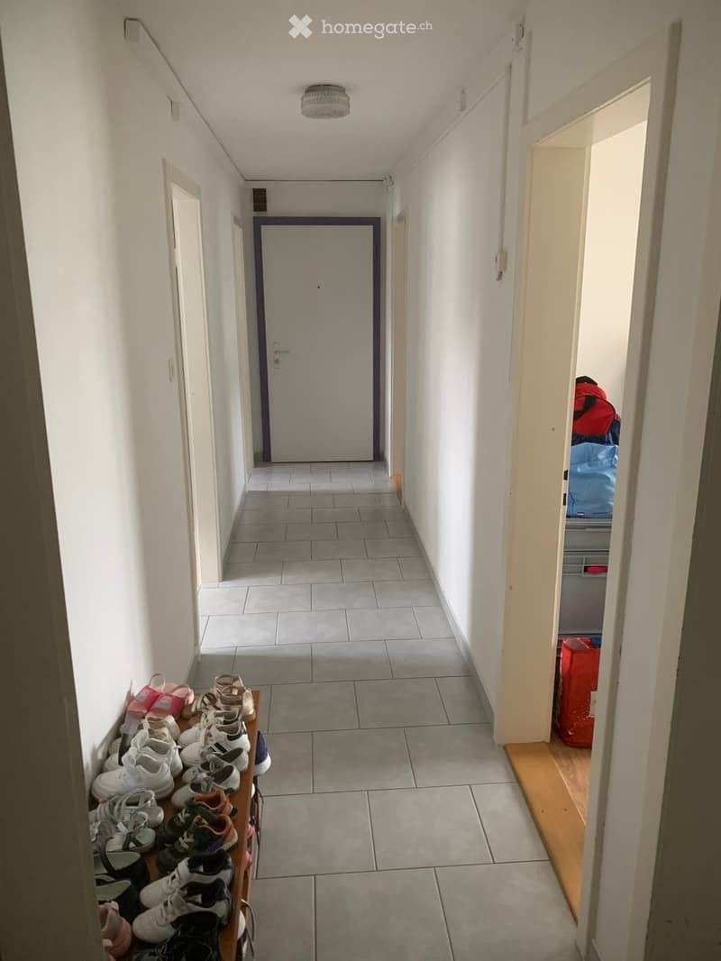 A narrow hallway with tiled flooring, white walls, and a ceiling light fixture. A shoe rack with multiple pairs of shoes is positioned on the left side, and a partially open door on the right reveals some storage items.